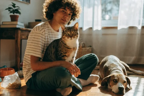 Young adult sitting on the floor with a cat in their lap and dog beside them, surrounded by pet supplies