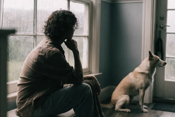 Person sitting by a window on a rainy day with a pet leash hanging by the door